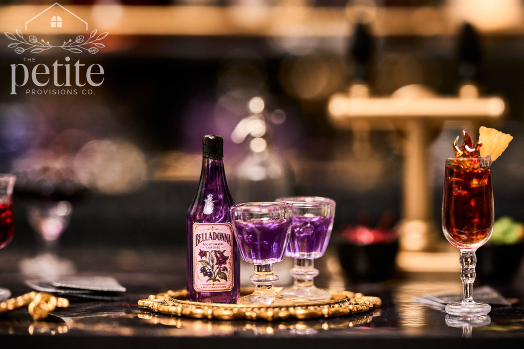A bottle of Belladonna Nightshade Cordial and two small glasses filled with purple liquid on a gold tray.