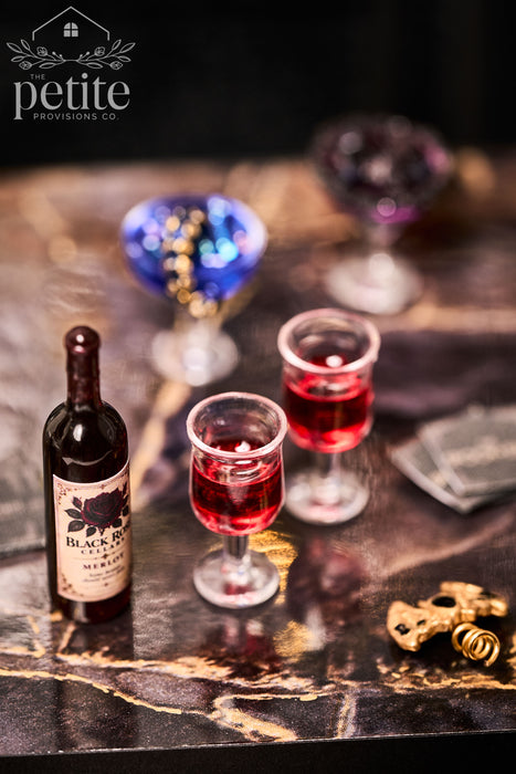 A bottle of Black Rose Cellars Merlot wine with two small glasses filled with red wine.