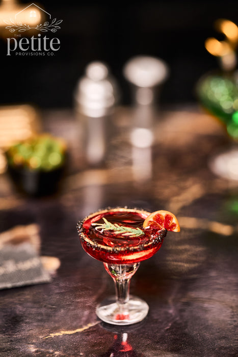 Handcrafted 1:12 scale Crimson Hour blood orange cocktail in a stemmed coupe with a black-and-gold rim, miniature blood orange slice, and rosemary garnish.