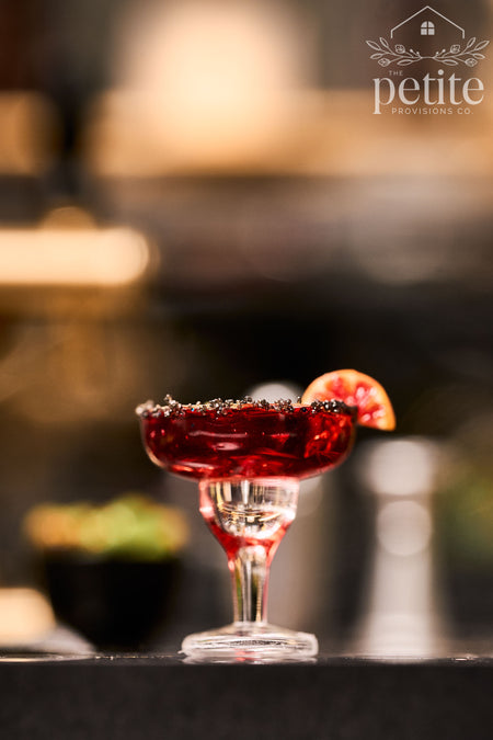 A dark red cocktail in a coupe glass with a salted rim and a citrus garnish, set on a dark bar counter with a blurred bar background. 