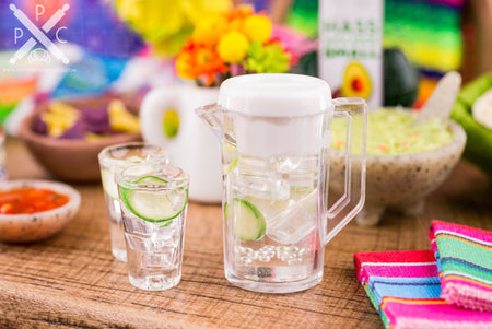 A dollhouse miniature replica of a refreshing drink. The pitcher of ice water is full of cucumber slices, and it comes with two matching glasses.