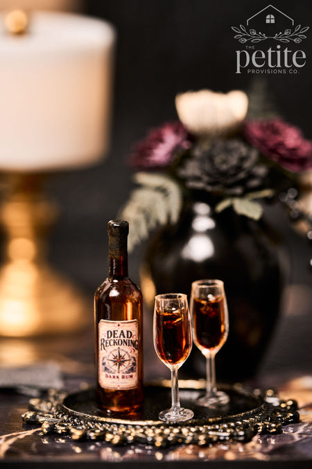 Close-up of a miniature Dead Reckoning Dark Rum bottle with a vintage compass rose label beside two slender glasses of amber resin rum on a decorative tray.
