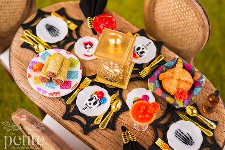 Overhead view of a 1:12 scale Día de los Muertos table setting for four with a gold lantern, handmade tamales, pan de muerto, miniature margaritas, sugar skull plates, and gold flatware.