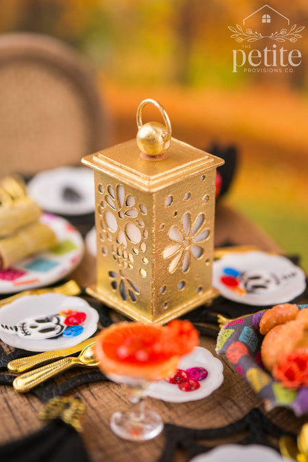 Handcrafted 1:12 scale gold Día de los Muertos lantern featuring intricate sugar skull and floral cutouts, displayed among miniature margaritas, traditional foods, and decorative plates.