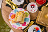 Three handmade 1:12 scale miniature tamales wrapped in corn husks on a colorful papel picado platter with an orange marigold, surrounded by Día de los Muertos dinnerware.