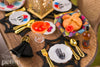 Close-up of a 1:12 scale Día de los Muertos table setting featuring a miniature marigold margarita, pan de muerto, gold flatware, sugar skull plates, and black spiderweb placemats.