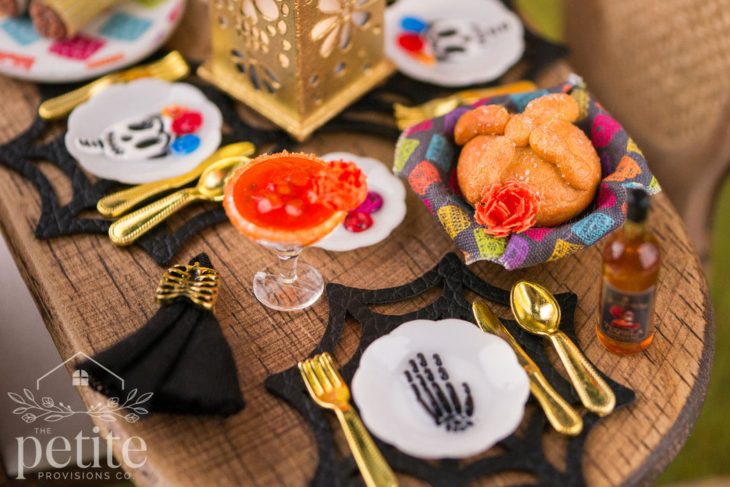 Close-up of a 1:12 scale Día de los Muertos table setting featuring a miniature marigold margarita, pan de muerto, gold flatware, sugar skull plates, and black spiderweb placemats.