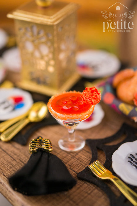 Handcrafted 1:12 scale Flor de Cempasúchil Margarita with an orange marigold garnish and colorful rim, displayed beside a black napkin with a gold ribcage ring and miniature gold flatware.