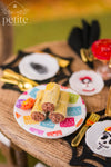 Close-up of three handmade 1:12 scale miniature tamales wrapped in textured corn husks and tied with strips of husk, displayed on a colorful papel picado platter with an orange marigold.