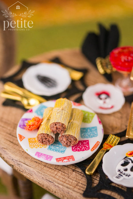 Close-up of three handmade 1:12 scale miniature tamales wrapped in textured corn husks and tied with strips of husk, displayed on a colorful papel picado platter with an orange marigold.