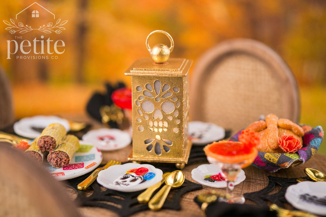 Handcrafted 1:12 scale gold sugar skull lantern surrounded by miniature tamales, pan de muerto, margaritas, decorative skull plates, and gold flatware in a Día de los Muertos table setting.