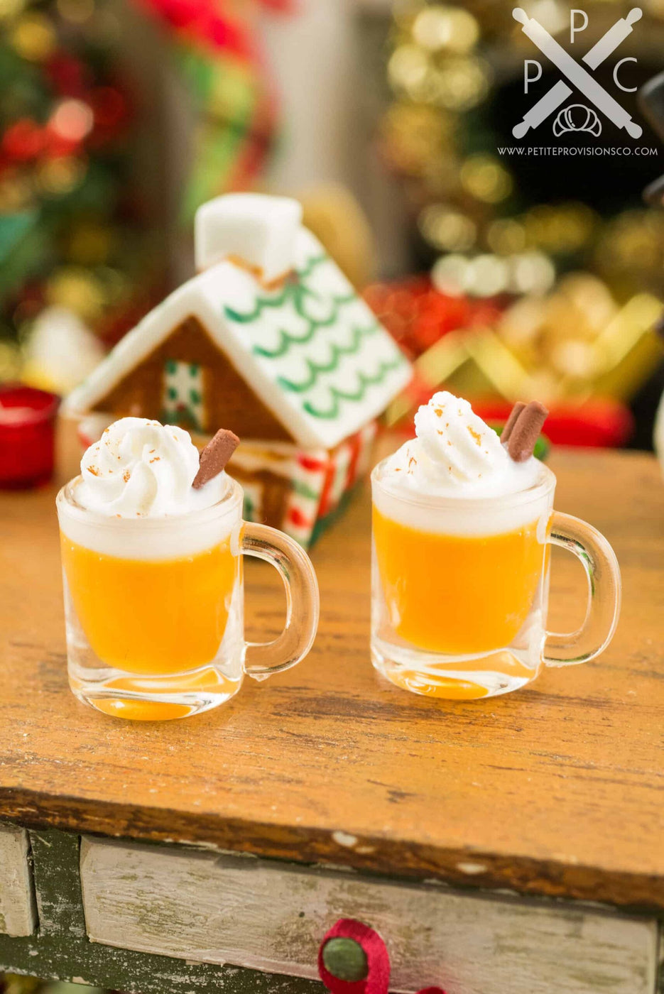 Two miniature mugs of hot buttered rum topped with whipped cream, styled with a tiny gingerbread house.