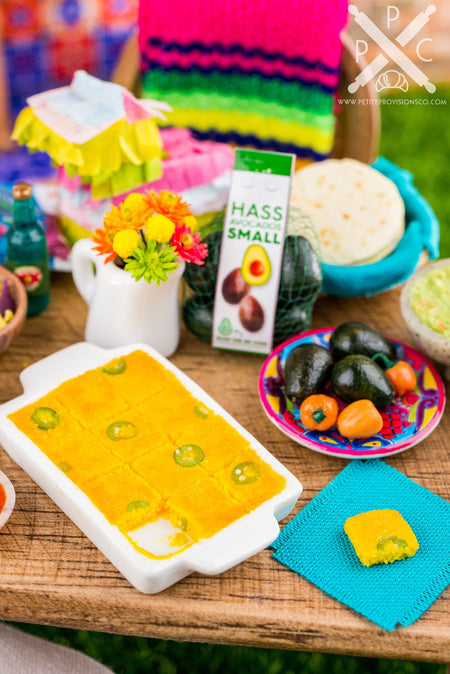 A dollhouse miniature white ceramic casserole dish filled with freshly baked jalapeño cornbread. A square has been removed from the cornbread, and a teal fabric napkin is placed on the side.