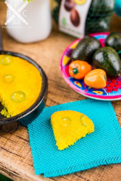 A miniature skillet made of ceramic and polymer clay, painted to look like a jalapeno cornbread skillet. The skillet is accompanied by a teal napkin with a slice of jalapeno cornbread.