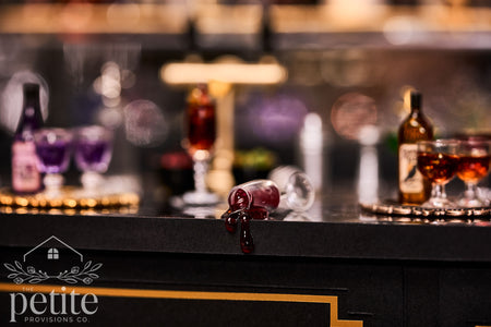 Handcrafted 1:12 scale miniature overturned wine glass spilling glossy dark red resin wine over the edge of a black Art Deco bar.