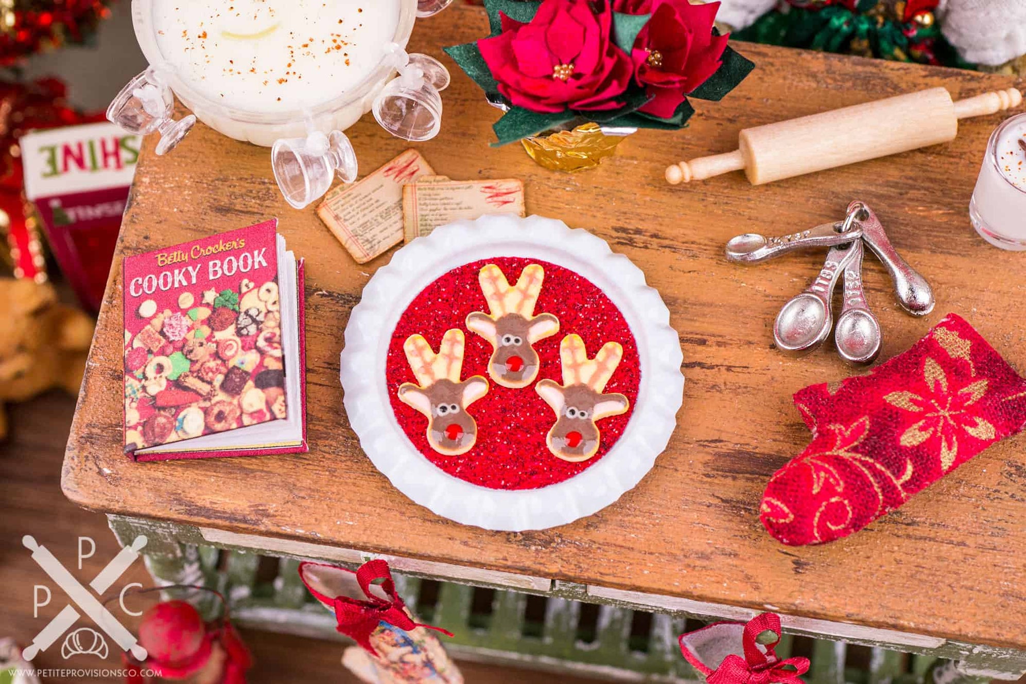 Miniature reindeer Christmas cookies on a red tray surrounded by tiny baking supplies and a vintage cookbook.
